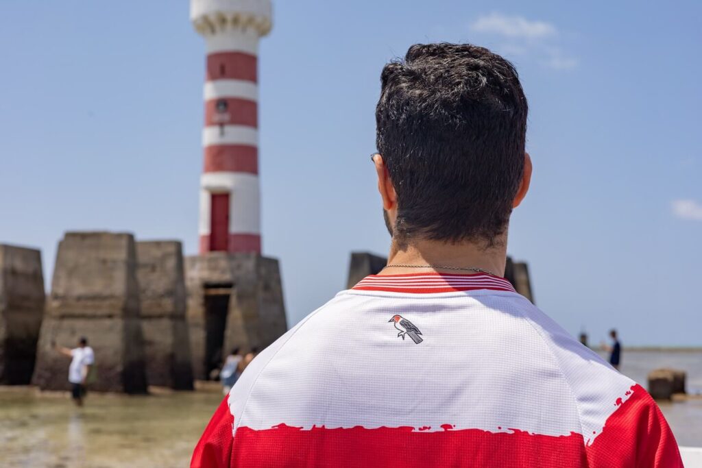 O CRB lançou oficialmente a sua terceira camisa para a temporada 2024-2025 em comemoração aos 112 anos do clube. Desenvolvida pela marca Regatas, o uniforme tem como destaque o design inspirado no Farol da Ponta Verde, um dos ícones de Maceió. Com predominância de branco e listras horizontais vermelhas em estilo “pintado”, a nova camisa reflete a tradição e a paixão regatiana. A peça também conta com detalhes especiais, como a frase “Alagoas, estrela radiosa”, do hino do estado, e o primeiro escudo do CRB em branco no peito. Esta edição comemorativa promete ser um sucesso entre os torcedores, reforçando o orgulho de ser regatiano. Disponível para compra, a nova camisa do CRB será usada durante a campanha do clube na Série B 2024.